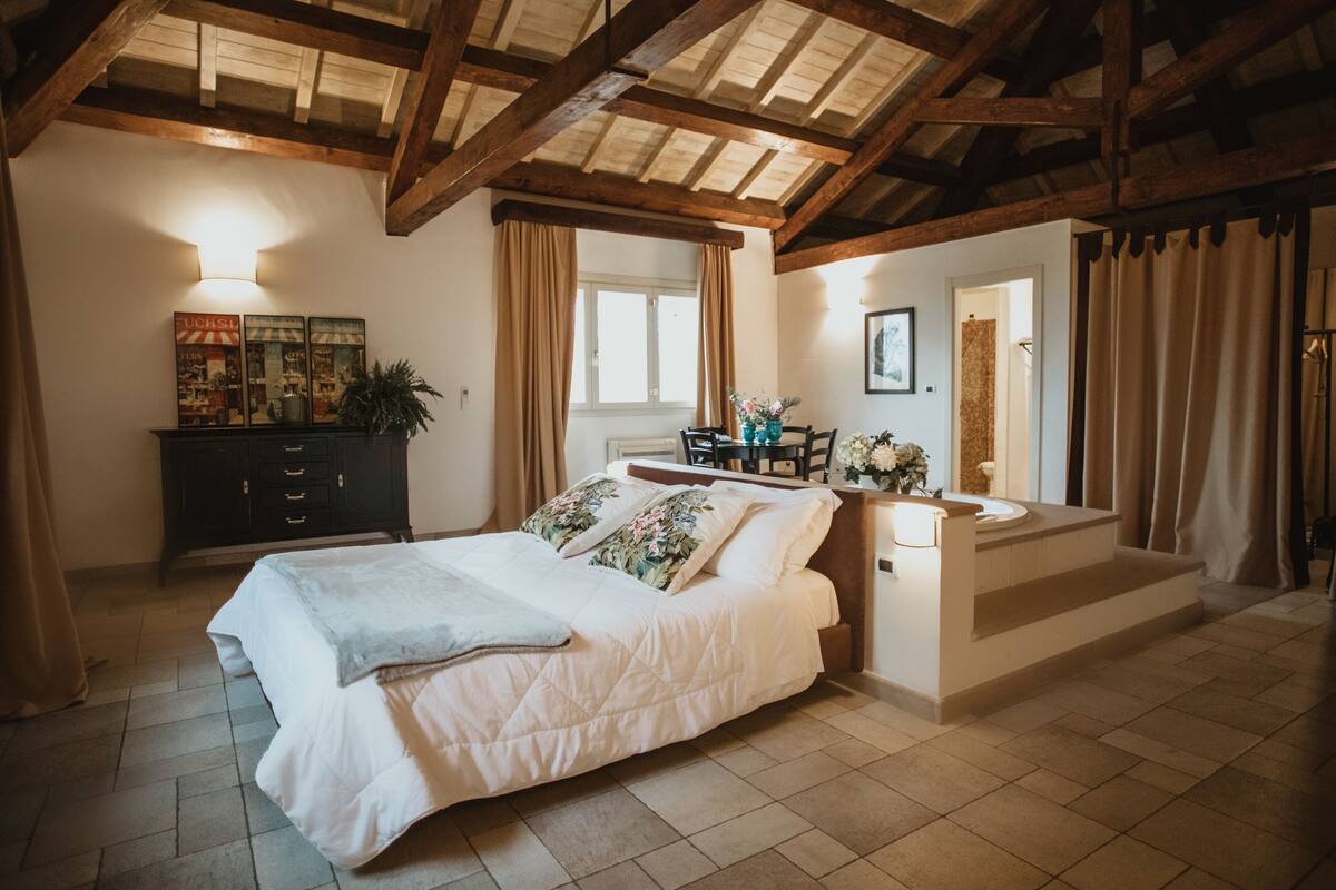 A bedroom with a large bed, a dresser, a vanity, and a window with curtains.