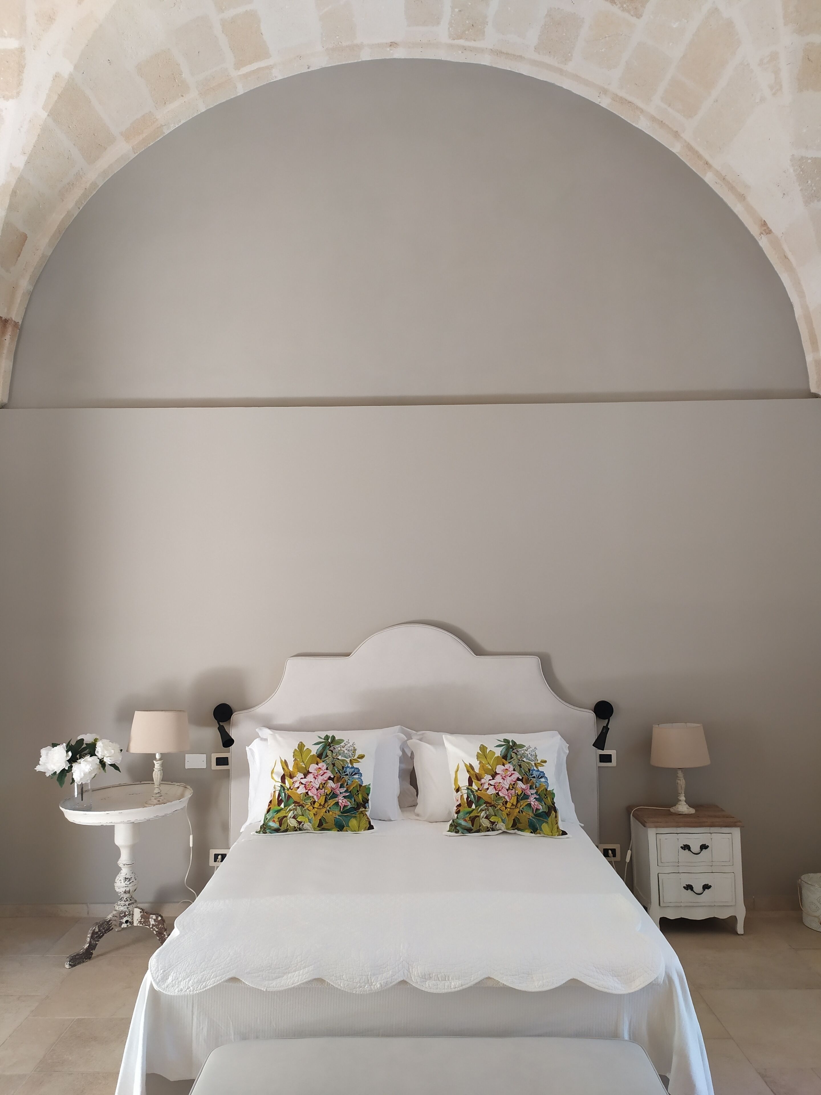 Un dormitorio con cama blanca, almohadas con motivos florales, una mesita de noche blanca con lámpara y un aparador blanco con lámpara.