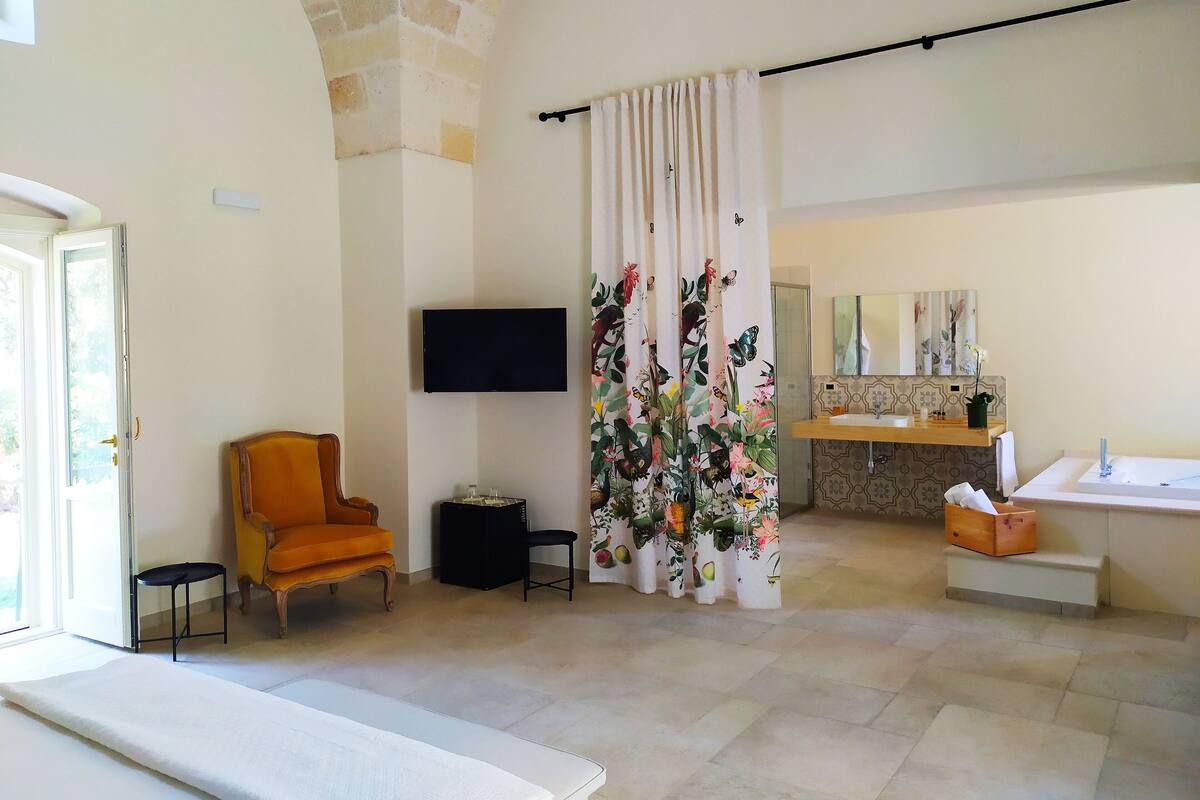 A spacious living room with a flat-screen TV, a comfortable orange armchair, a small side table, and a large window.