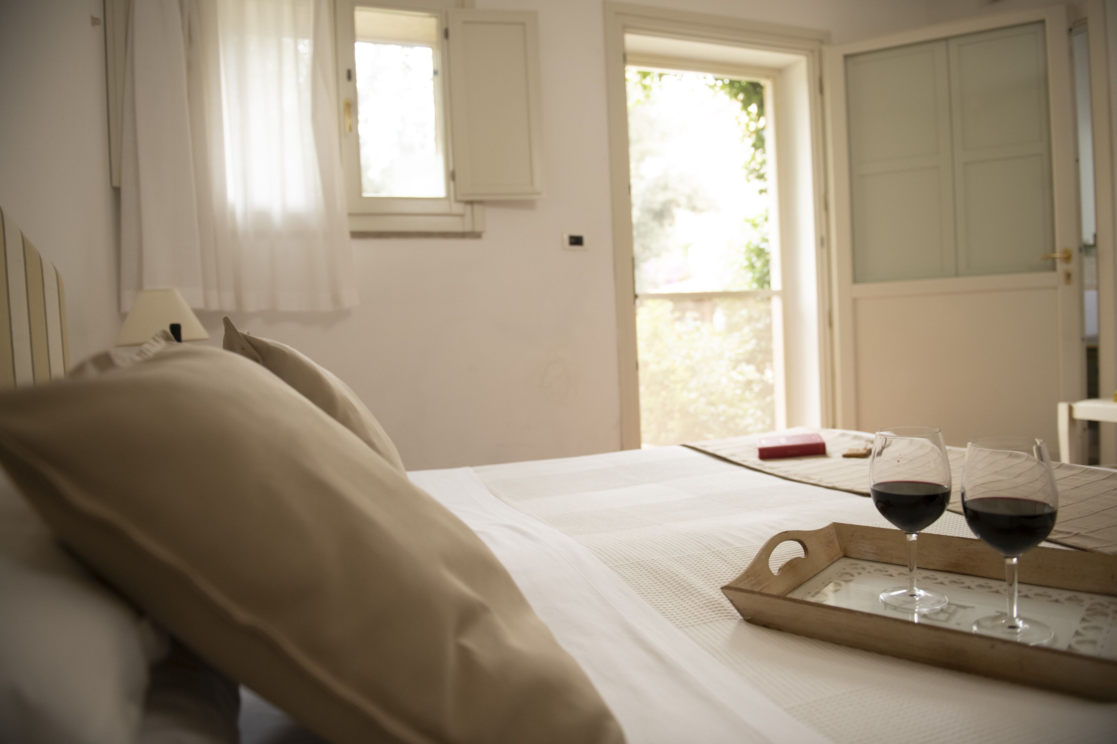 A bedroom with a bed, a window, and a tray with two glasses of red wine.