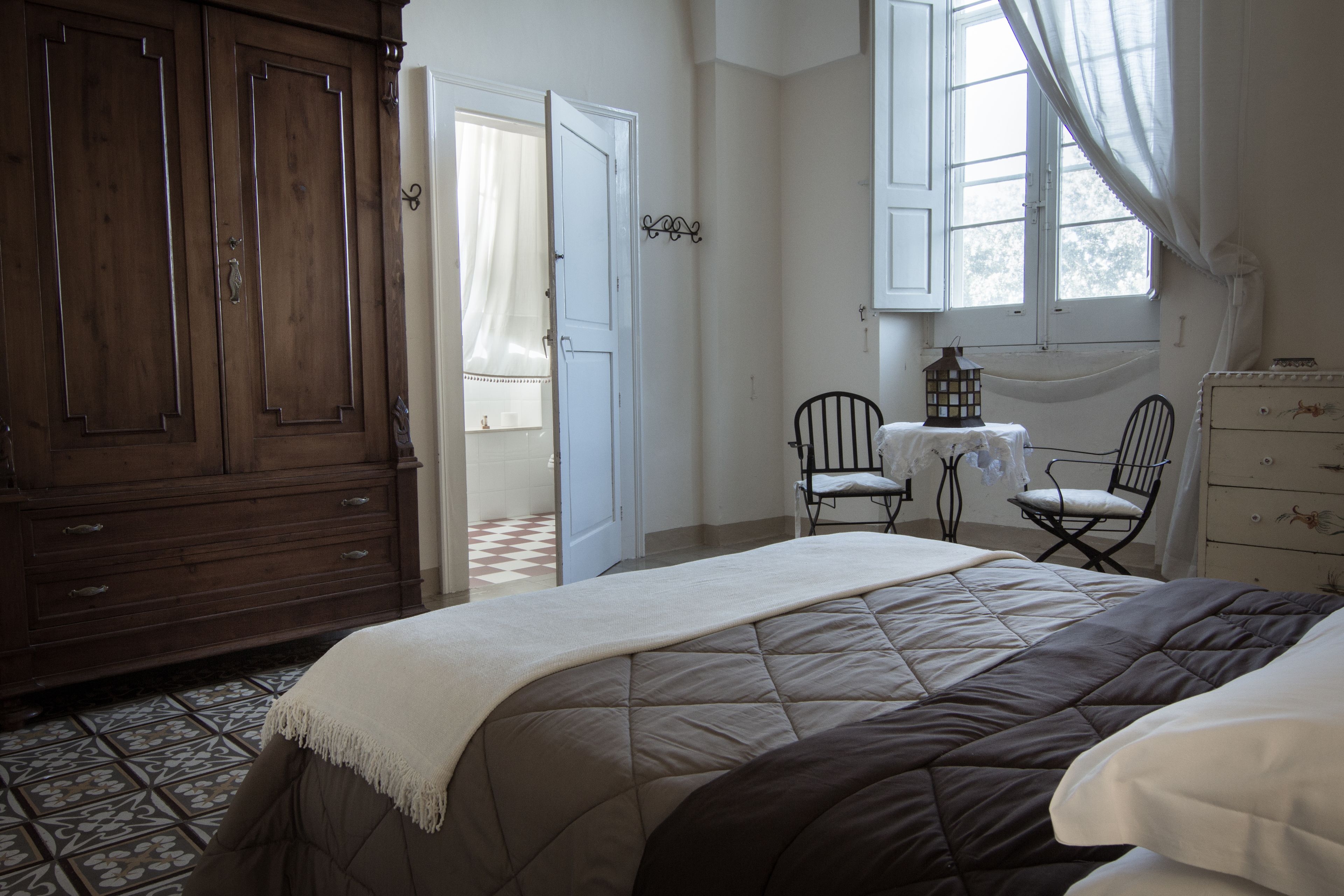 Une chambre à coucher avec un grand lit, une armoire en bois, une petite table avec des chaises et une fenêtre avec des rideaux.