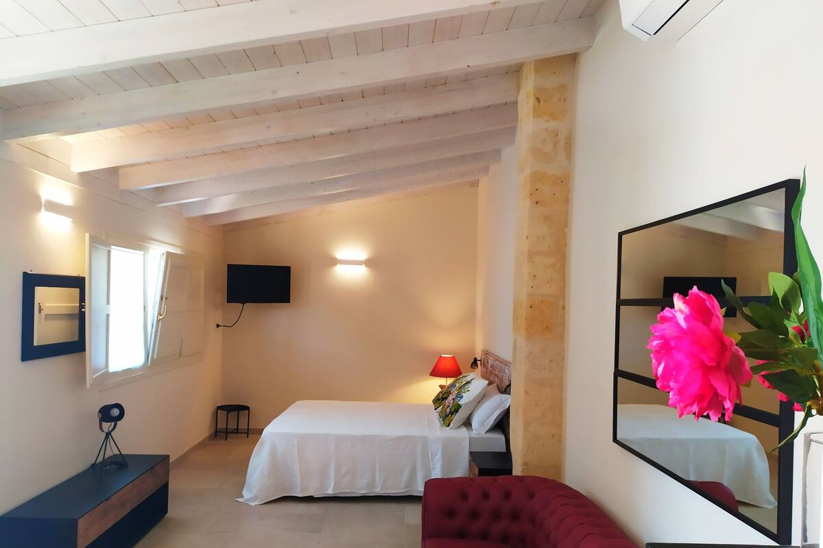 A bedroom with a bed, a red sofa, a TV, a mirror, and a window.