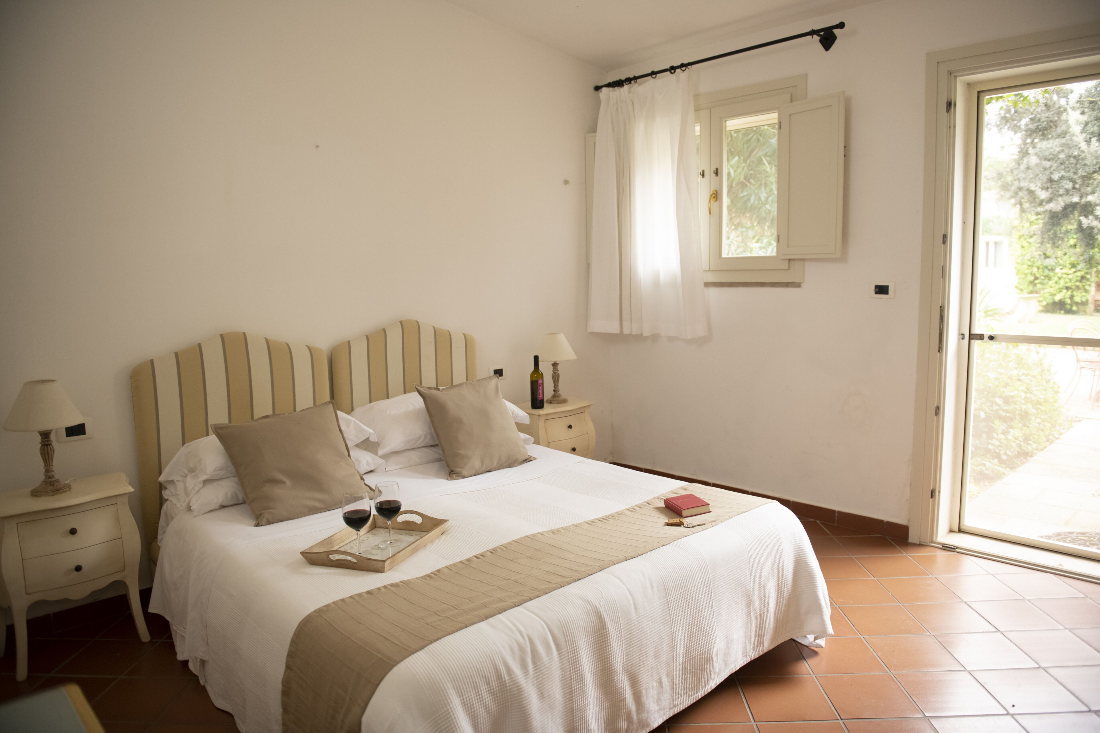 A neatly made bed with a tray of wine glasses and a book, a bedside lamp, and a window with curtains.