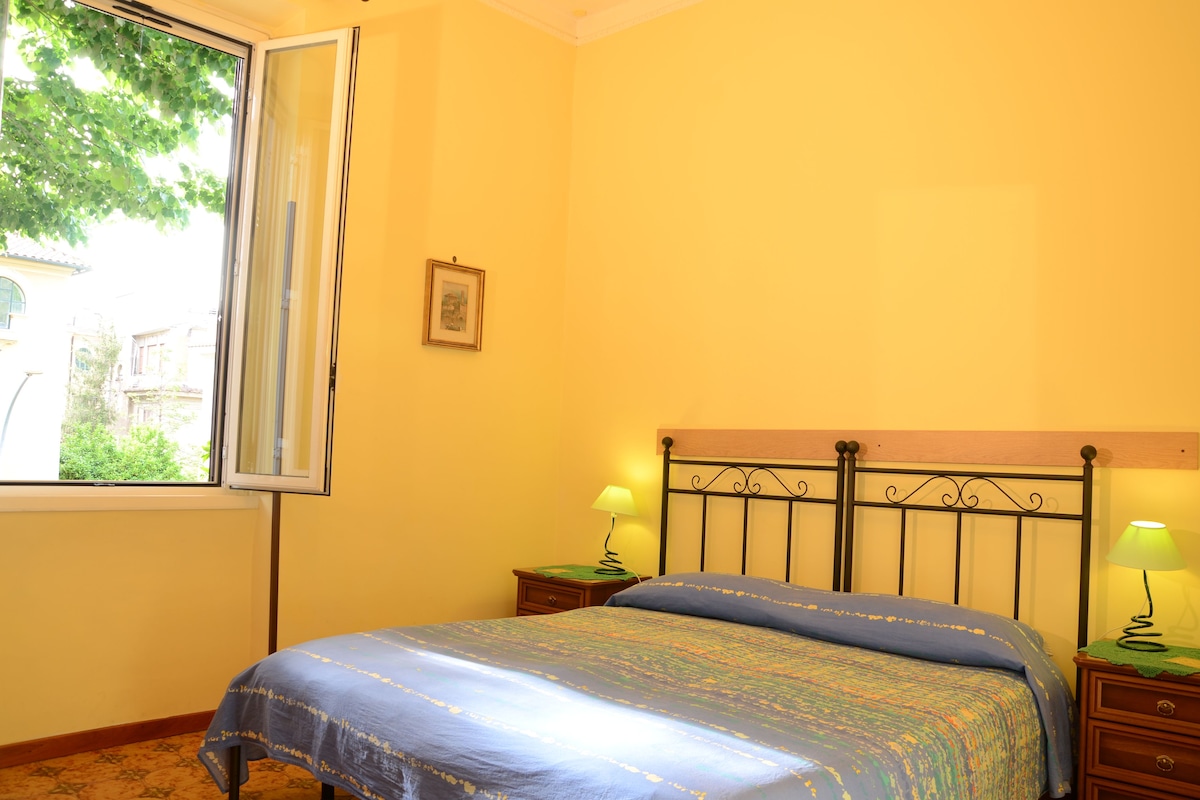 Une chambre à coucher avec un lit, deux tables de chevet avec des lampes, une fenêtre donnant sur un paysage verdoyant et un tableau encadré au mur.