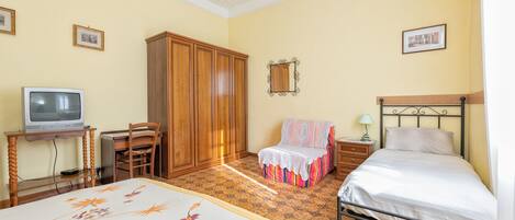 Un dormitorio con una cama, una silla, un escritorio con televisor, un armario y una mesita de noche con lámpara.
