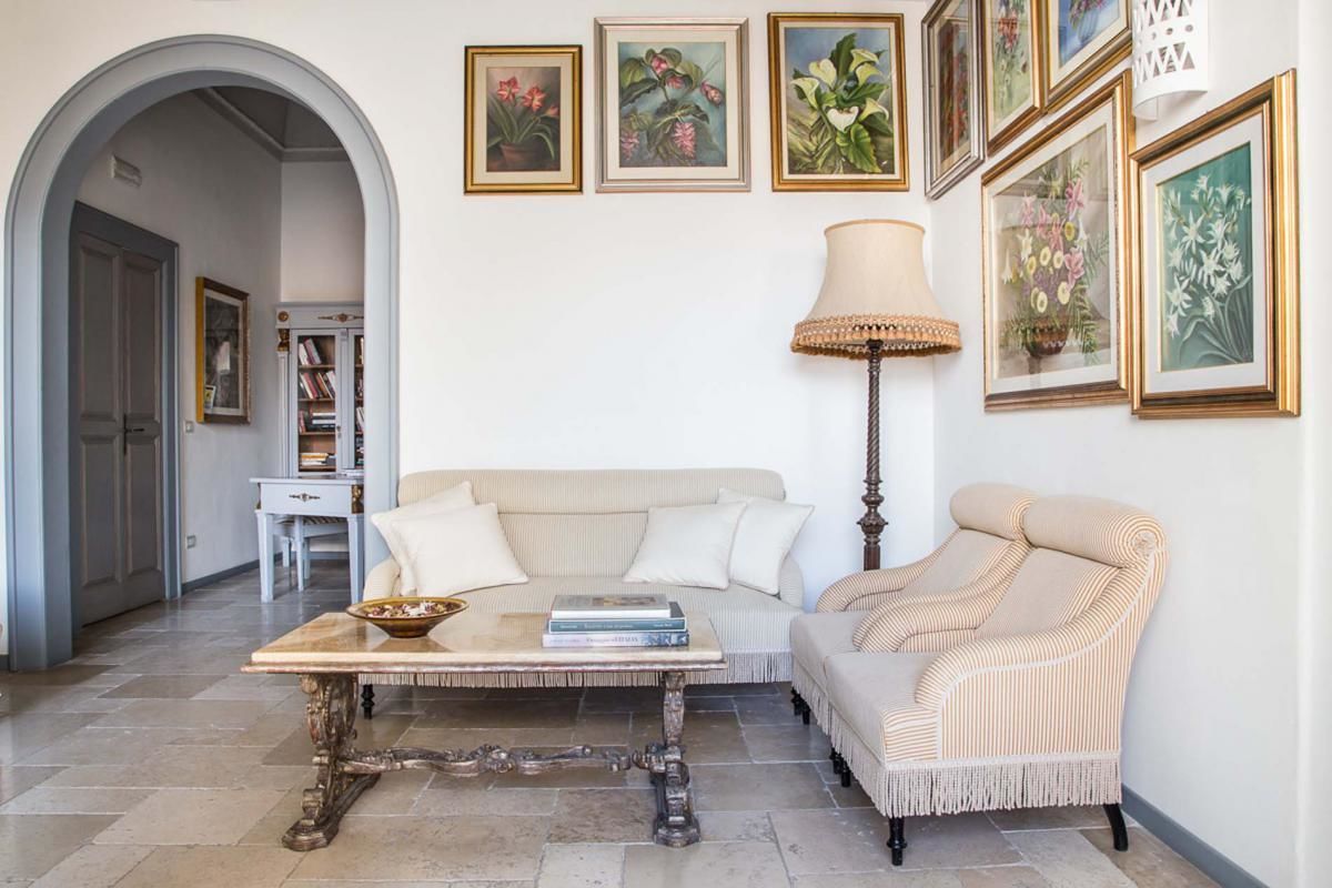 A living room with a sofa, armchair, coffee table, and a lamp. There are framed pictures on the wall and a bookshelf in the background.