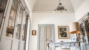 A room with a white ceiling, a chandelier, a desk with a lamp, and a bookshelf. - Palazzo Angelelli (Gallipoli)