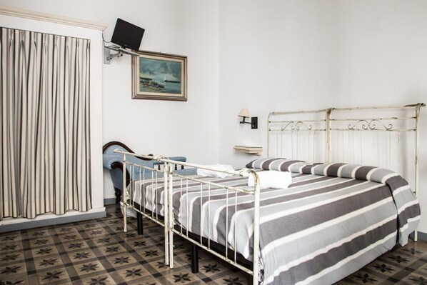 A bedroom with a metal bed frame, striped bedding, a painting on the wall, a television, and a window with curtains. - Palazzo Angelelli (Gallipoli)