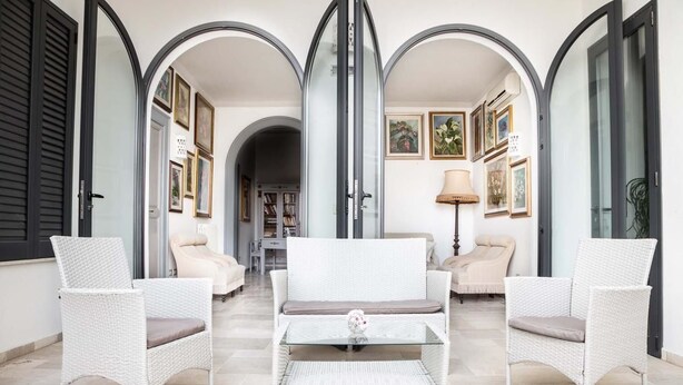 A room with a white wicker sofa set, a glass coffee table, and a lamp. There are framed pictures on the walls and a door with a glass panel. - Palazzo Angelelli (Gallipoli)