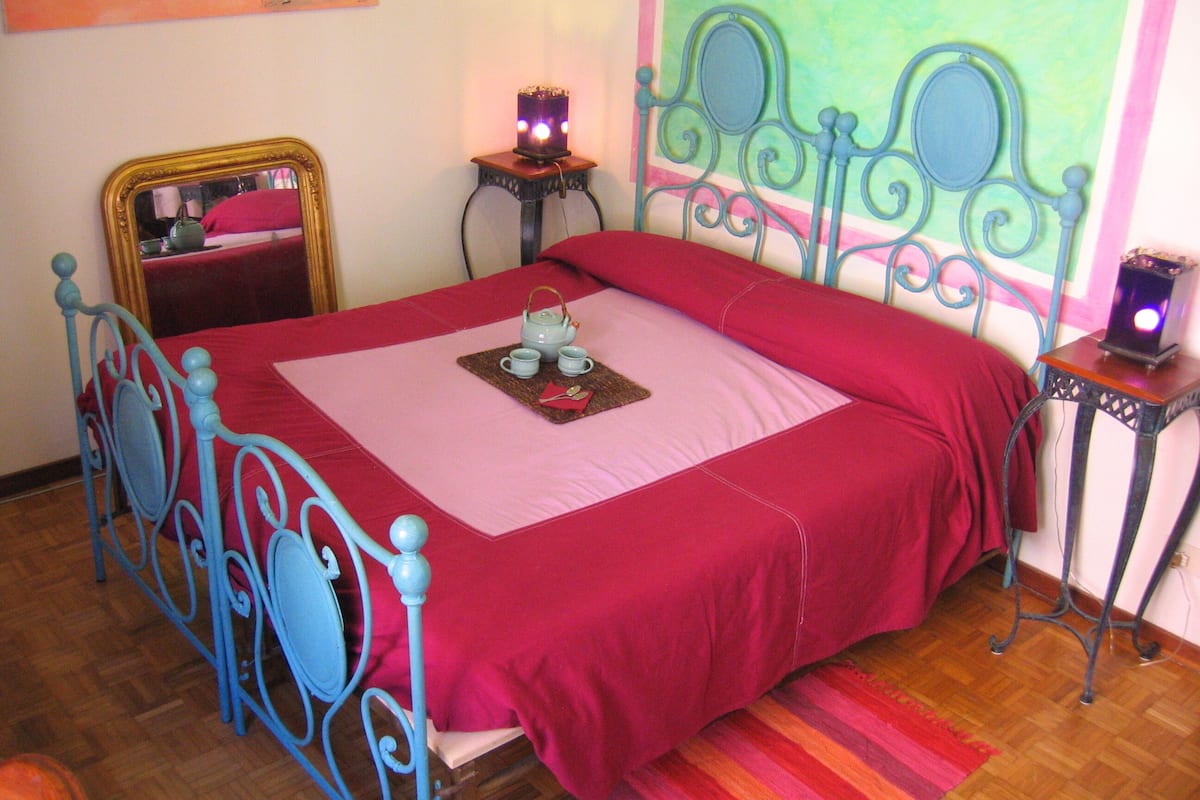 Un dormitorio con estructura de cama metálica, una colcha roja y una mesita de noche con lámpara.