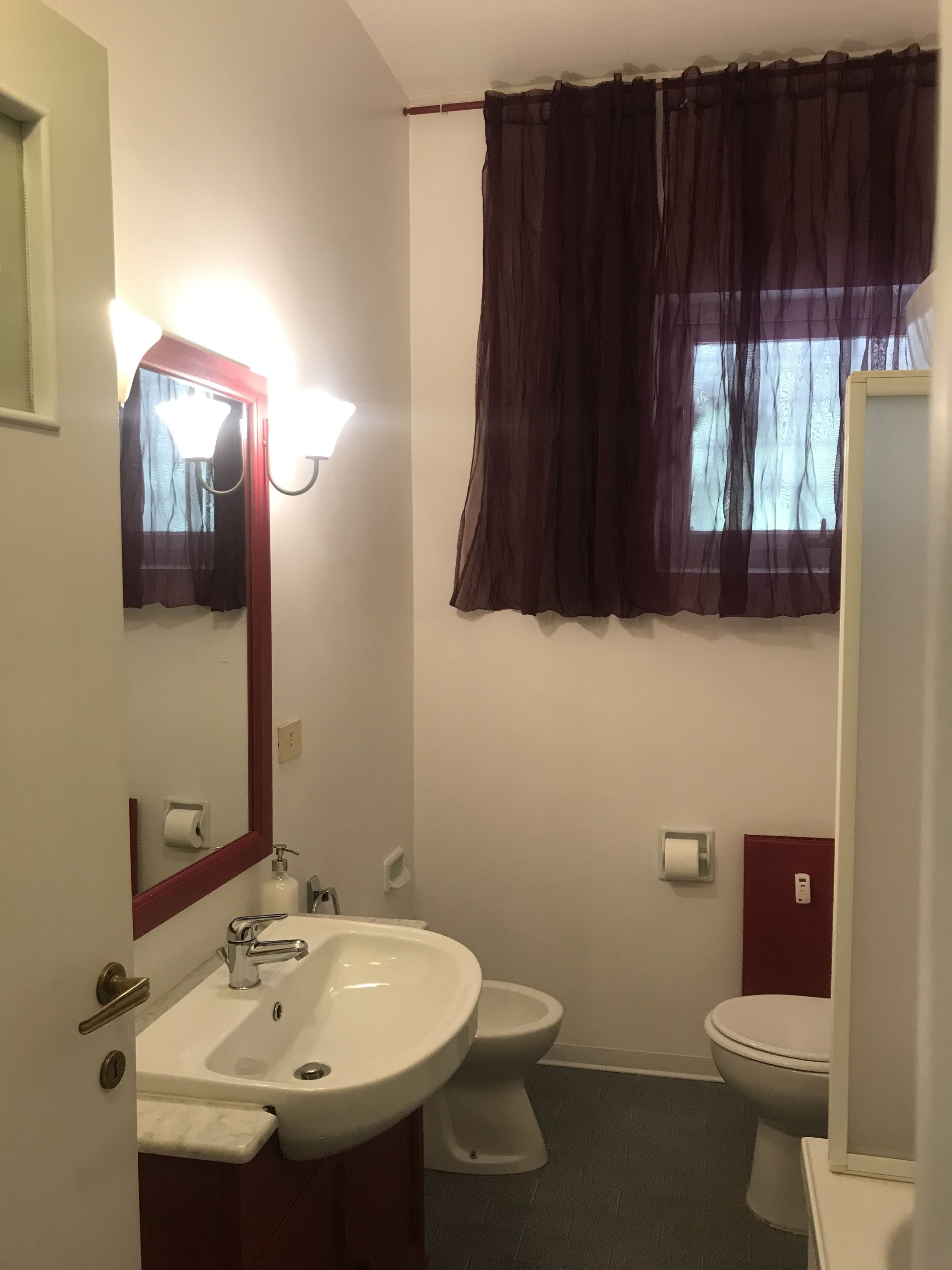 A bathroom with a white sink, two toilets, a mirror with a light, and a window with sheer curtains.