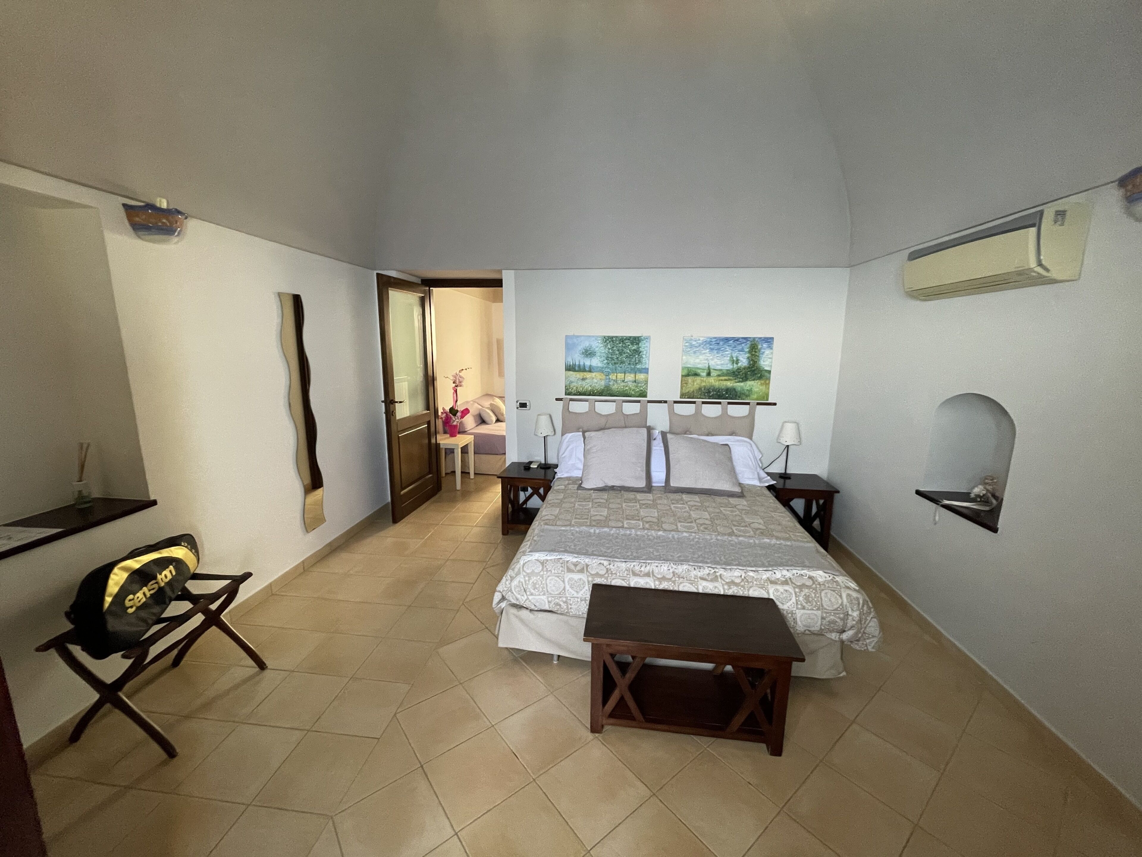A bedroom with a bed, bedside tables, a chair, a small table with a vase, and a mirror on the wall.