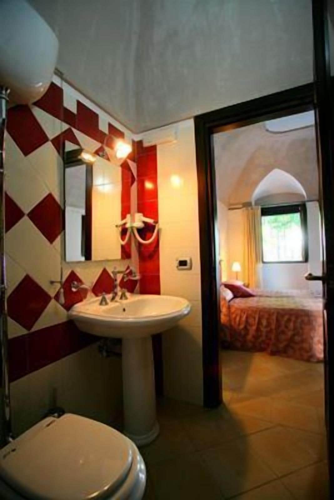 A bathroom with a sink, mirror, and toilet. There is a bed visible through an open door in the adjacent room.