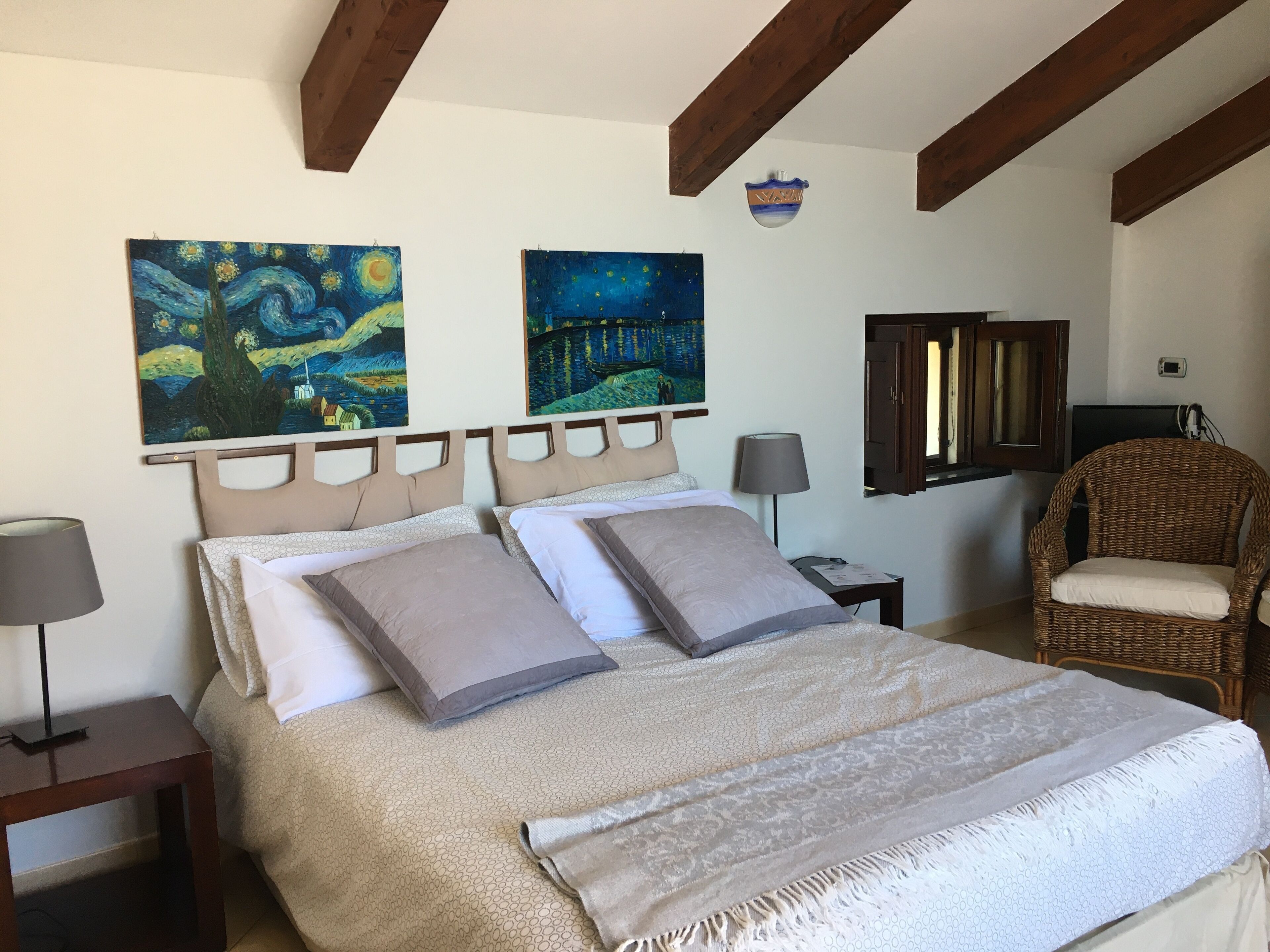 A bedroom with a bed, two paintings, a wicker chair, a lamp, and a small table.