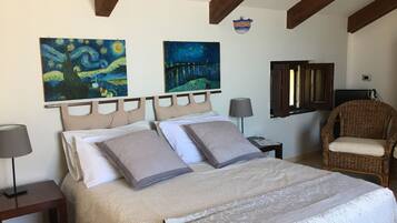 A bedroom with a bed, two paintings, a wicker chair, a lamp, and a small table.