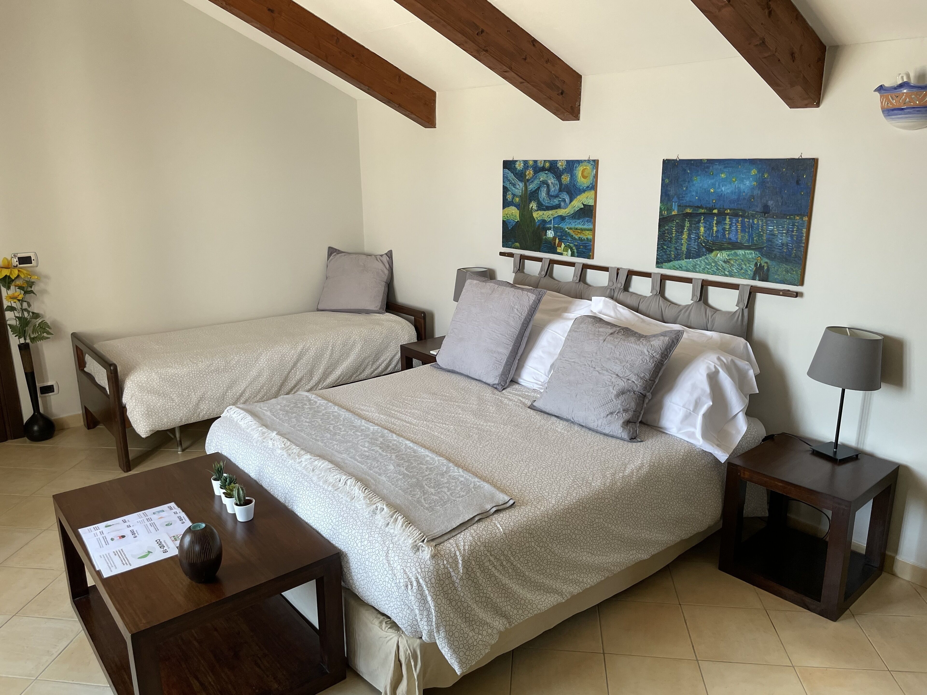 Un lit double avec une tête de lit, deux tables de chevet, une petite table d’appoint, une lampe, un vase avec des fleurs, une petite table avec un menu et deux tableaux accrochés au mur.