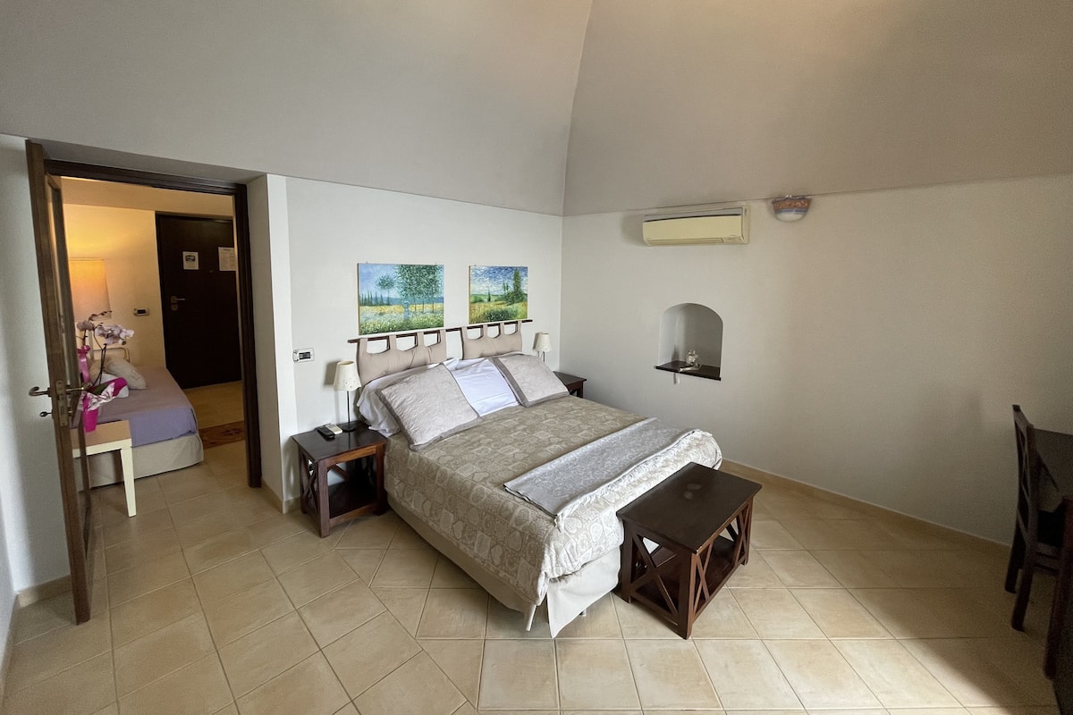 Ein Schlafzimmer mit Bett, Nachttischen, einem kleinen Holzbank und einem an der Wand montierten Regal. Eine Tür führt in einen anderen Raum.