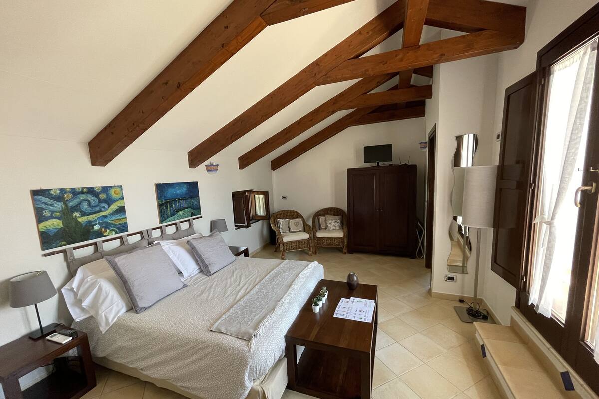 Ein Schlafzimmer mit einem großen Bett, zwei Gemälden, einer Holzbalkendecke, einem Kleiderschrank, einem Fernseher und einem Fenster.