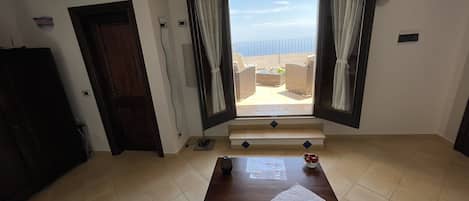 A room with a bed, a window with curtains, a wooden door, and a view of an outdoor balcony.