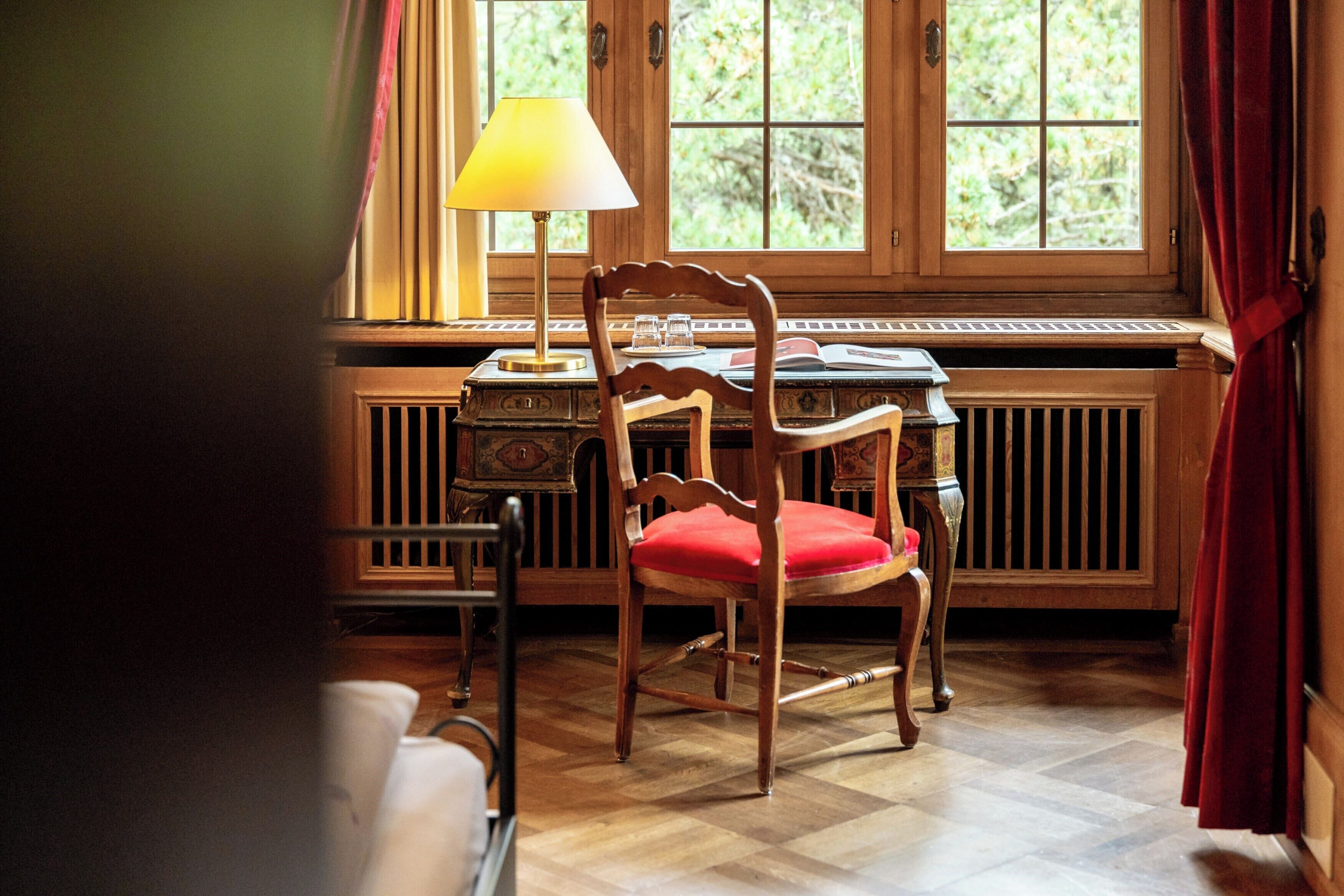 A wooden chair with a red cushion, a table with a lamp, and a window with curtains.