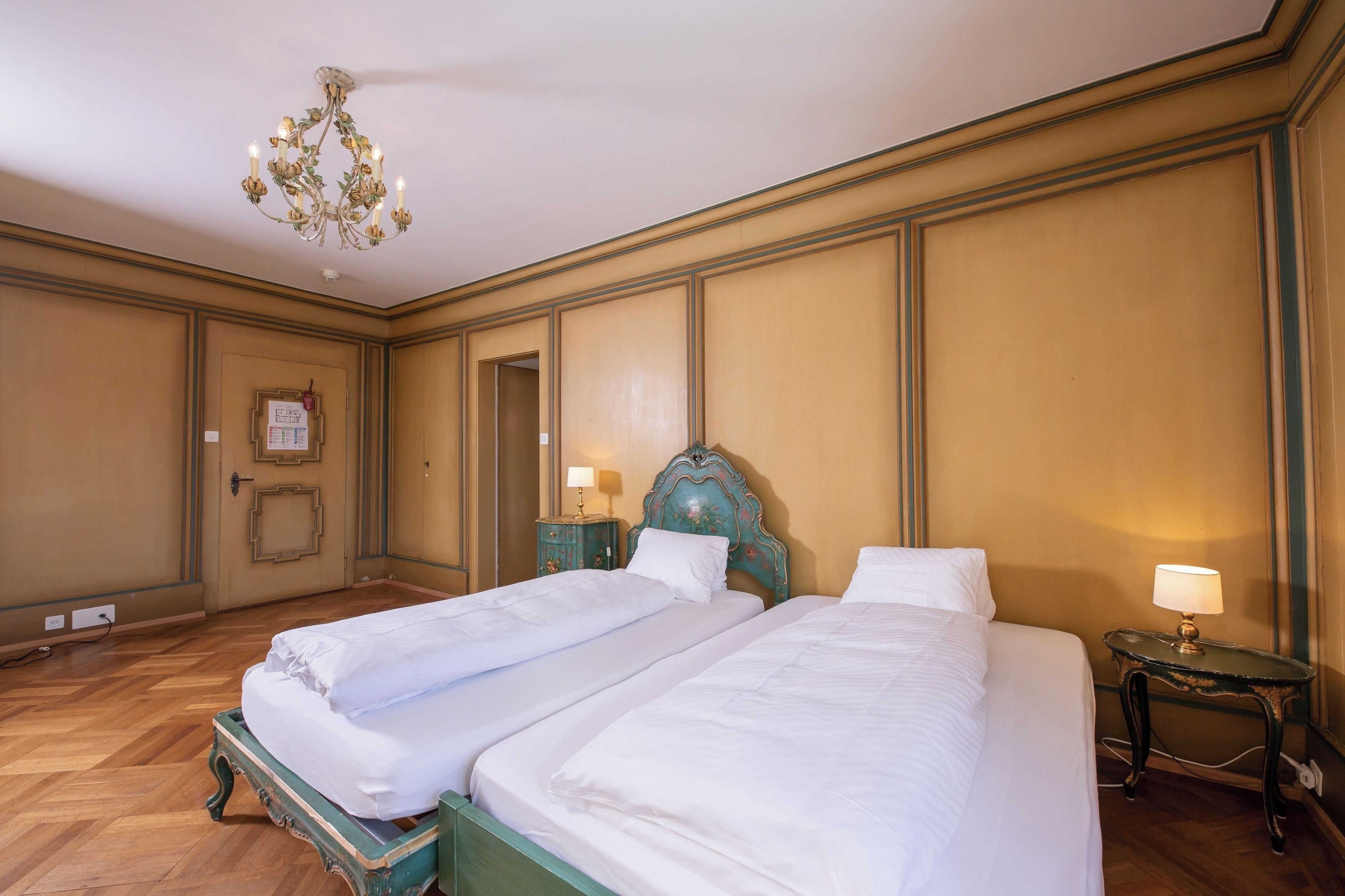 Ein Hotelzimmer mit zwei Betten, einer Kandelaber, einem kleinen Tisch mit Lampe und einem gerahmten Bild an der Wand.