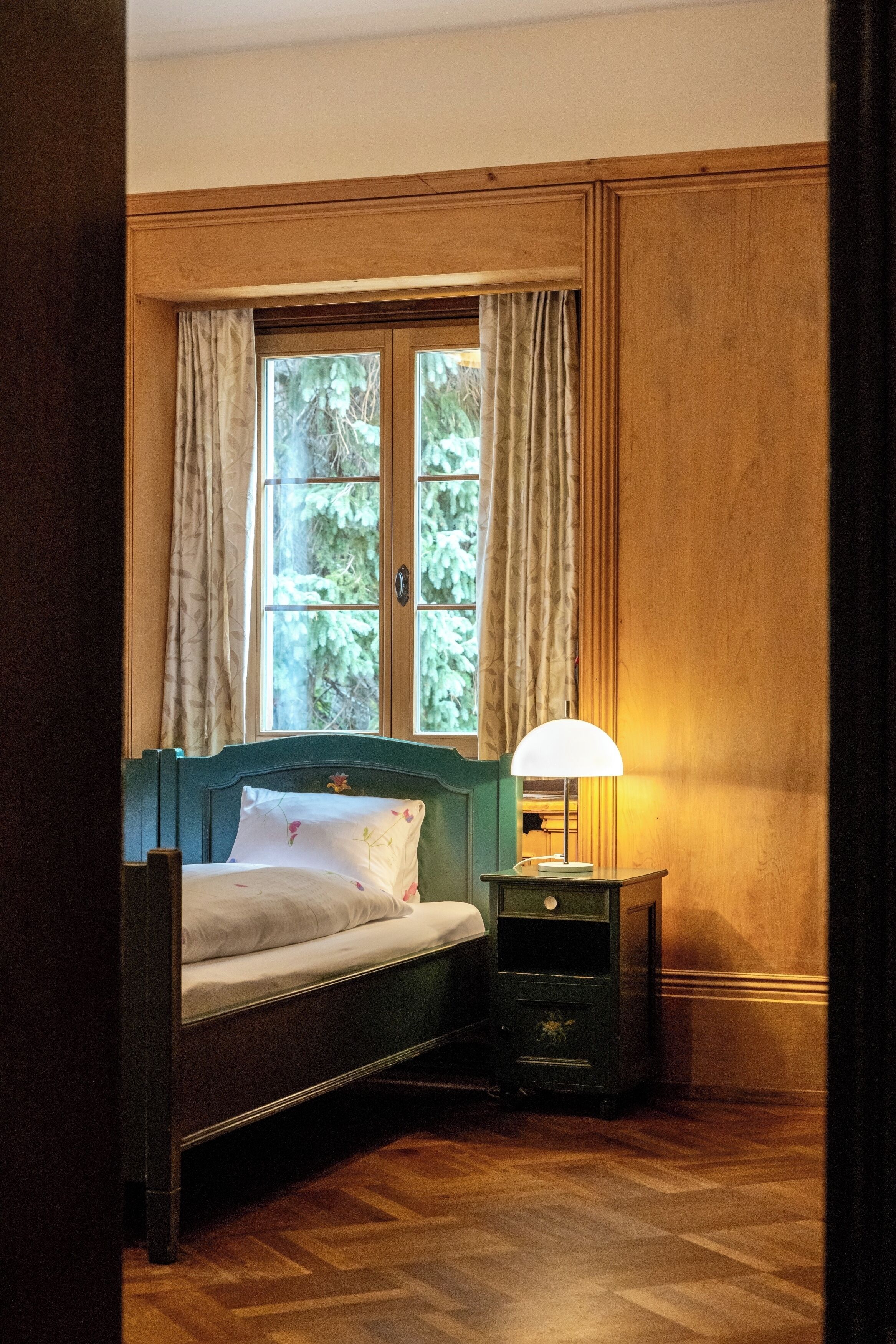 A bedroom with a wooden bed, a nightstand with a lamp, and a window with curtains.