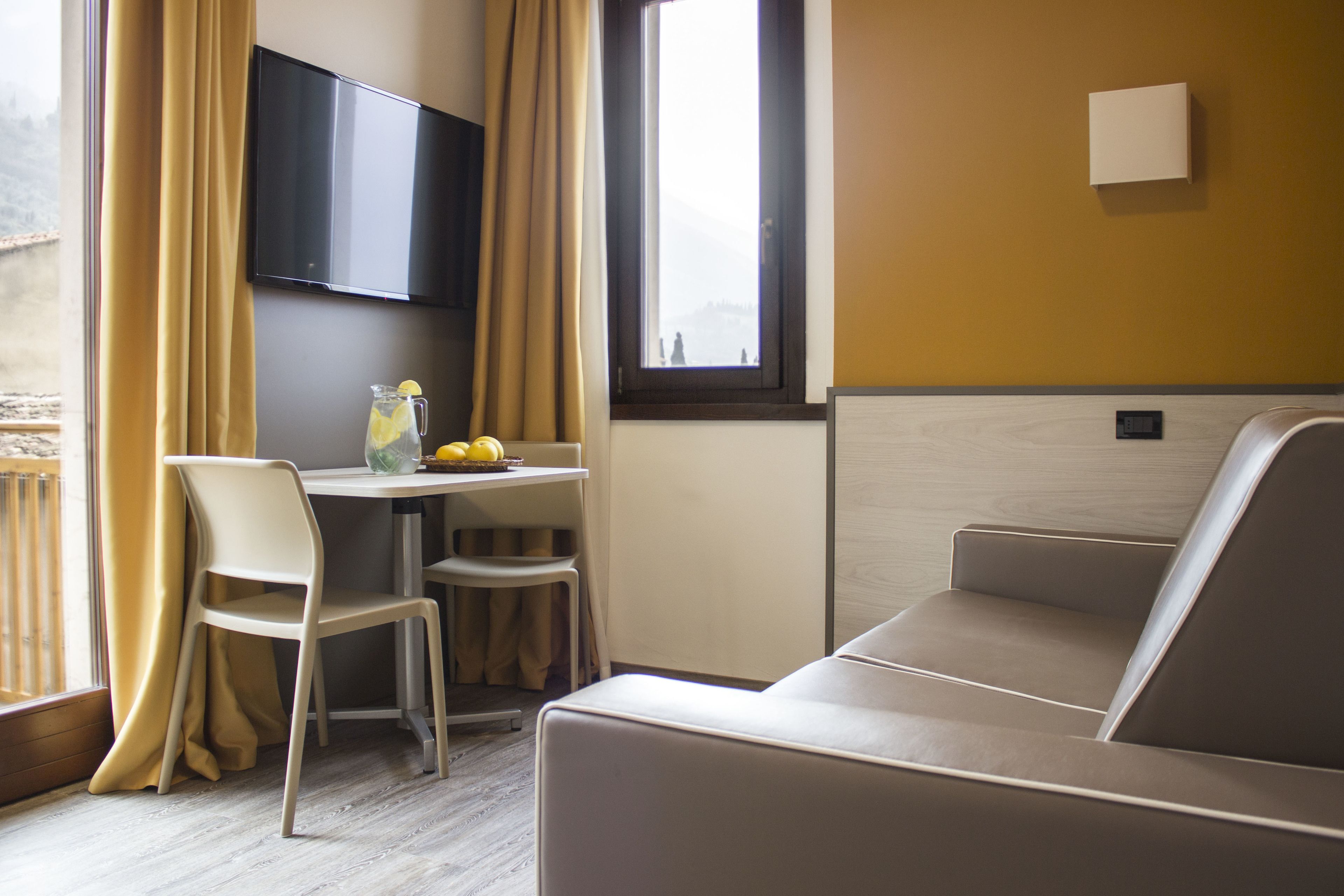 Una habitación moderna con sofá, una mesa con jarra de agua y limones, televisor montado en la pared y ventana con cortinas.