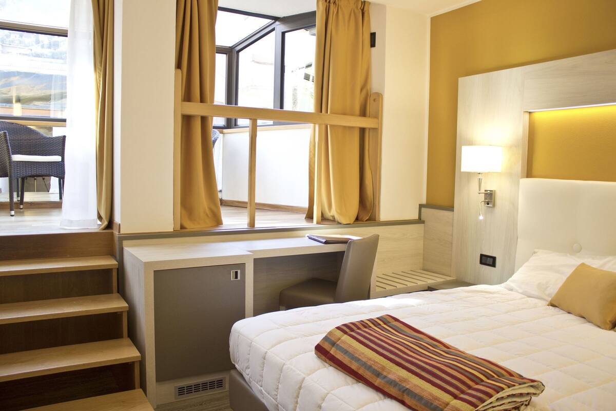 A hotel room with a bed, desk, chair, and a view of the outdoors.