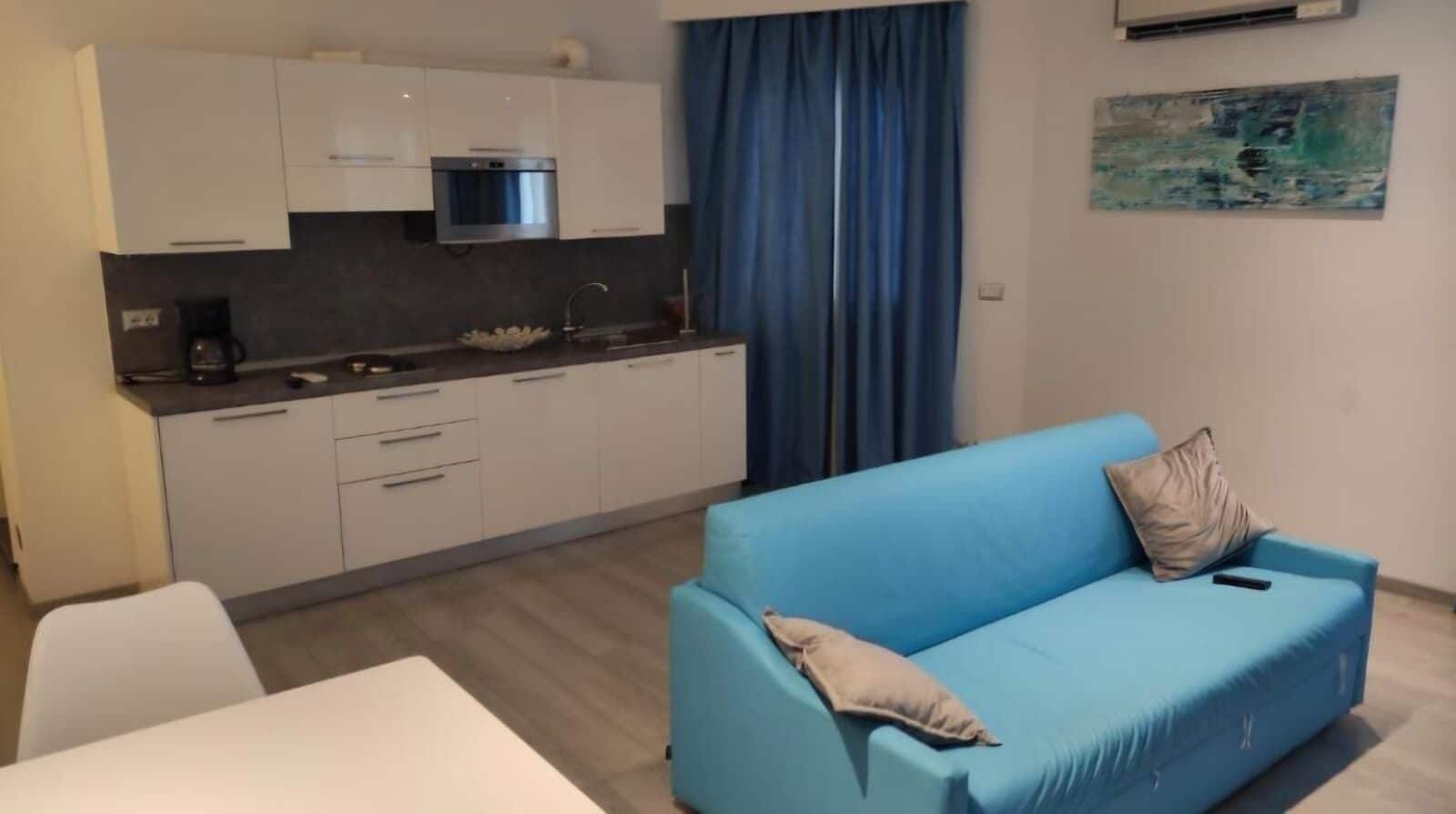 A modern kitchen with white cabinets, a built-in oven, and a coffee maker. There is a blue sofa with a cushion and a painting on the wall.