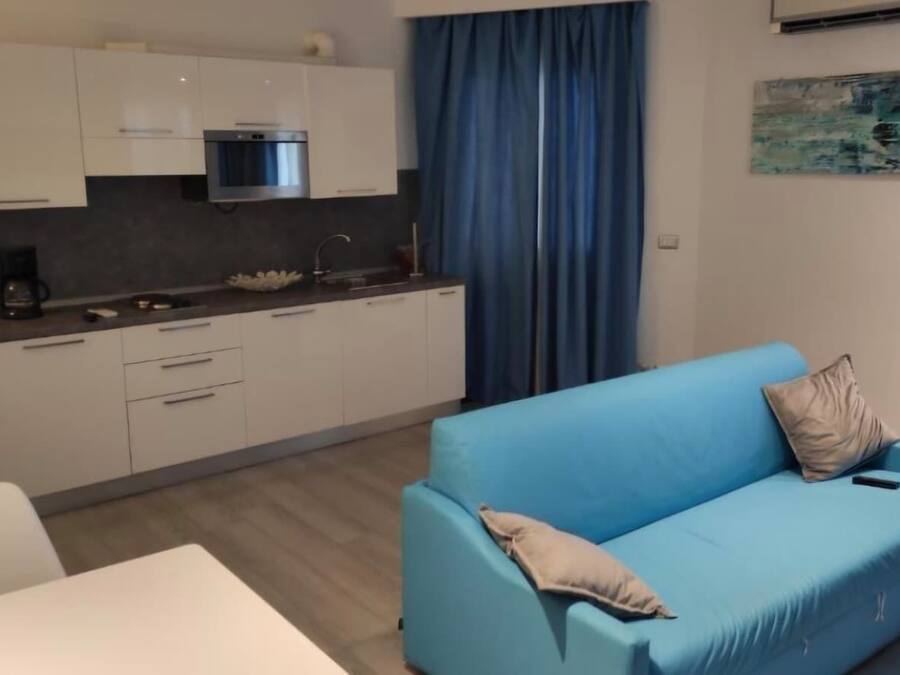 A modern kitchen with white cabinets, a built-in oven, and a coffee maker. There is a blue sofa with a cushion and a painting on the wall.