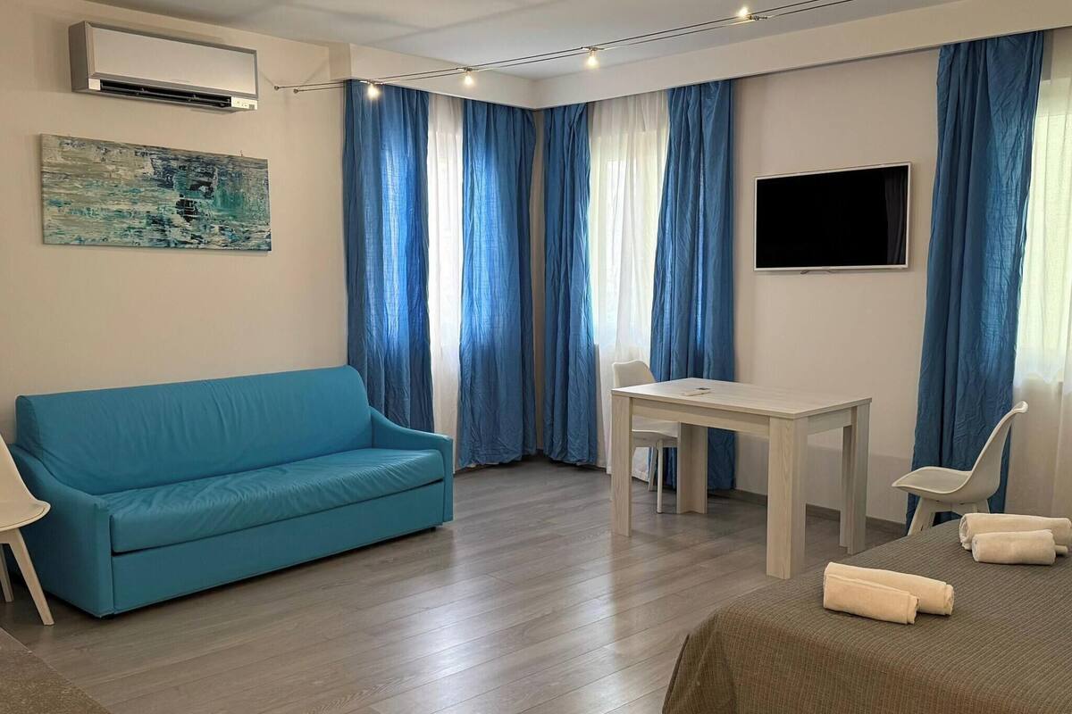 A room with a blue sofa, a small table, a bed, a TV, and a window with blue curtains.