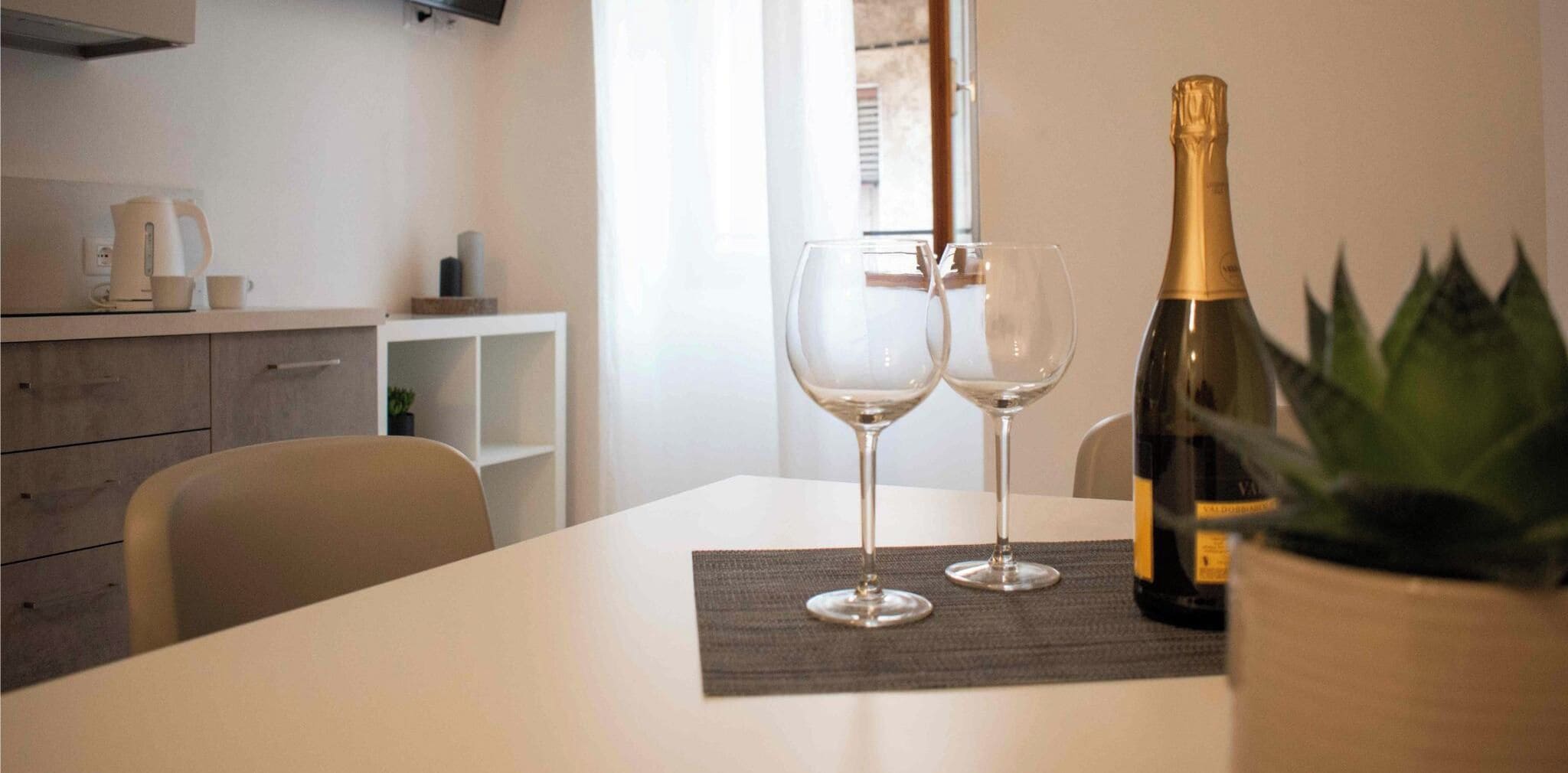 A dining table with two wine glasses and a bottle of champagne, a potted plant, and a kitchen area in the background.