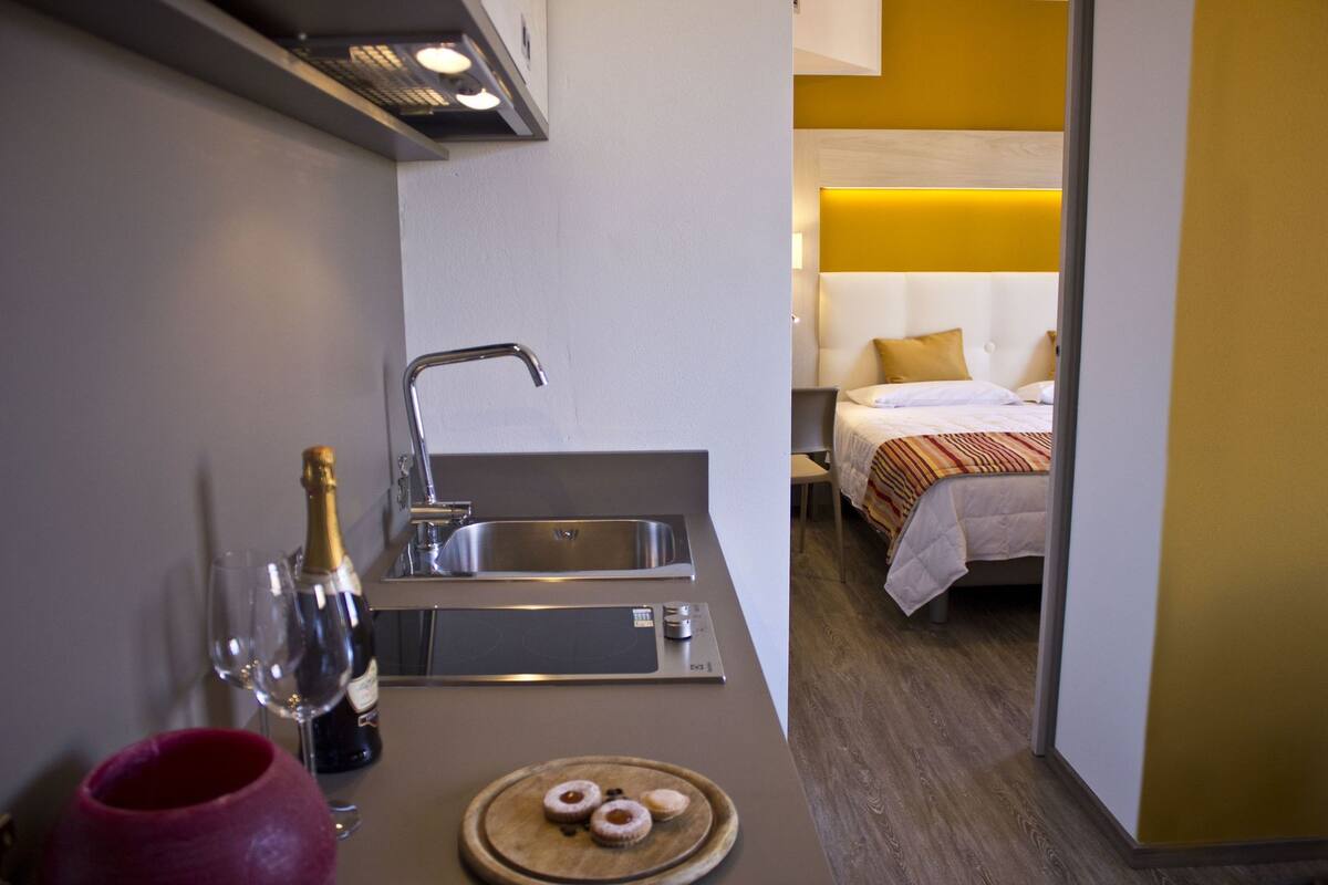 A compact hotel room with a kitchenette, a bed, and a bedside table.