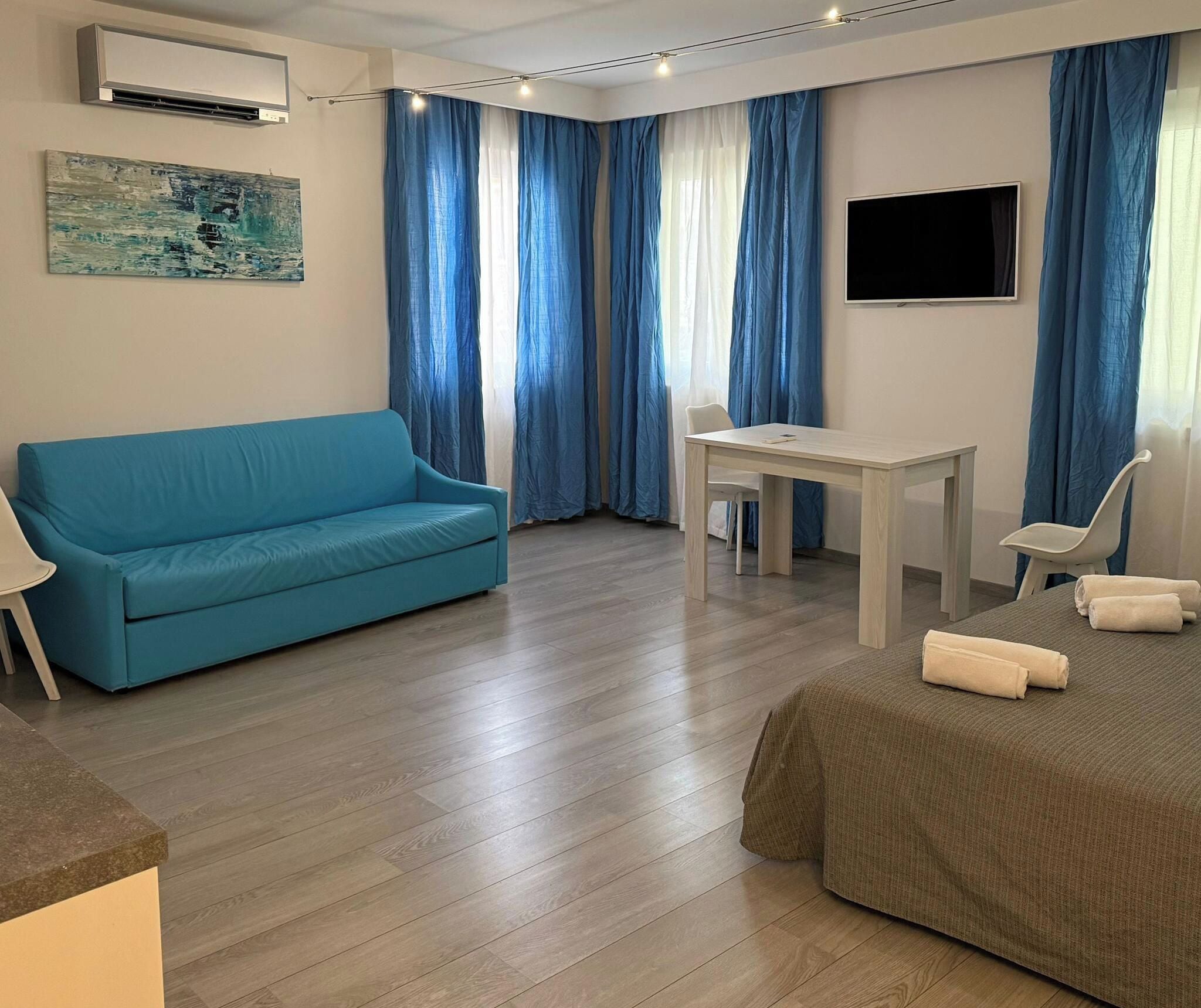 A room with a blue sofa, a small table, a TV, and a bed with towels.