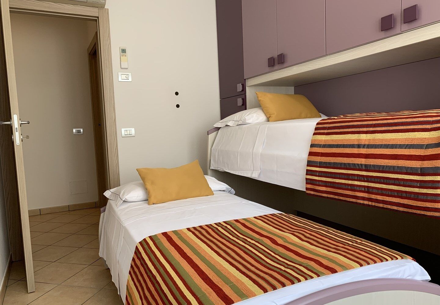 A room with two beds, one with a striped blanket and the other with a plain white bedspread. There are two pillows on the beds, one yellow and one white. The room has a door, a light switch, and a thermostat on the wall.