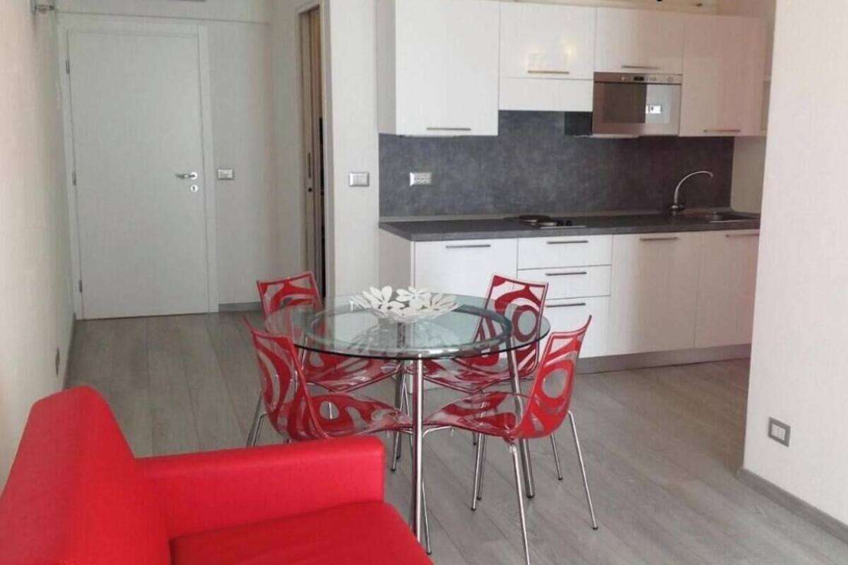 A compact kitchen with white cabinets, a black countertop, and a glass dining table with red chairs.