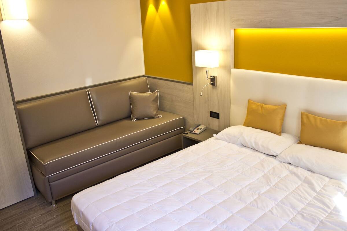 A hotel room with a bed, a sofa, a nightstand, and a wall-mounted lamp.