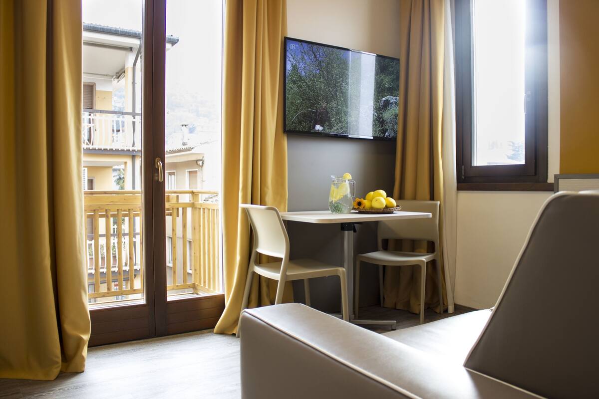 A room with a sofa, a table with a pitcher and lemons, a TV mounted on the wall, and a balcony with a railing.