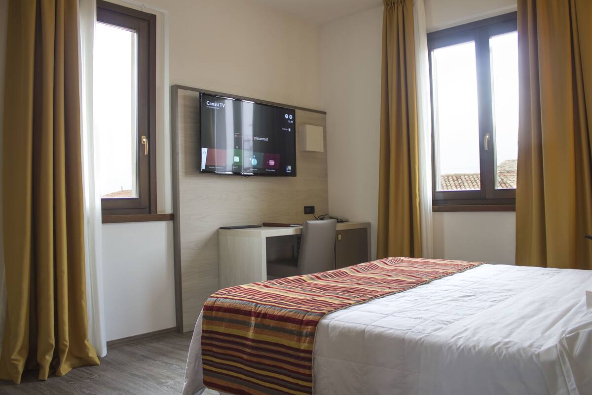 A hotel room with a bed, a desk, a TV mounted on the wall, and curtains on the windows.