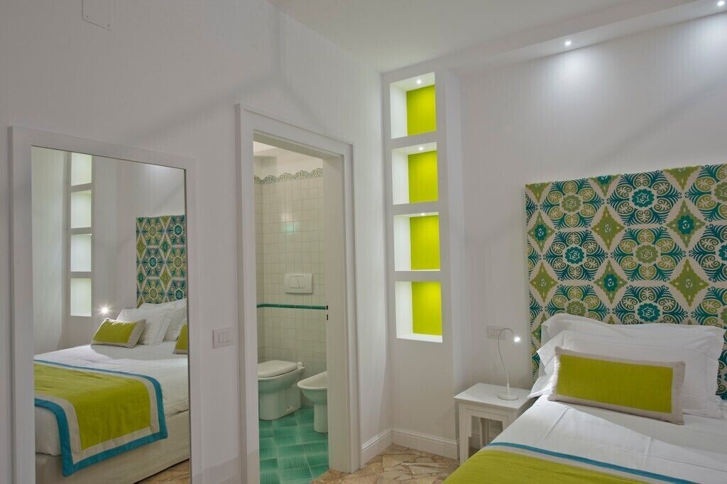 A hotel room with a bed, a bedside table, a mirror, a window, and a bathroom with a toilet and sink.