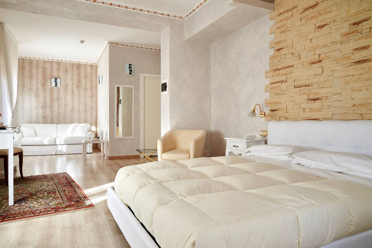 Una habitación de hotel moderna con una cama grande, una zona de estar con un sofá y sillón, una mesita pequeña y una alfombra decorativa.