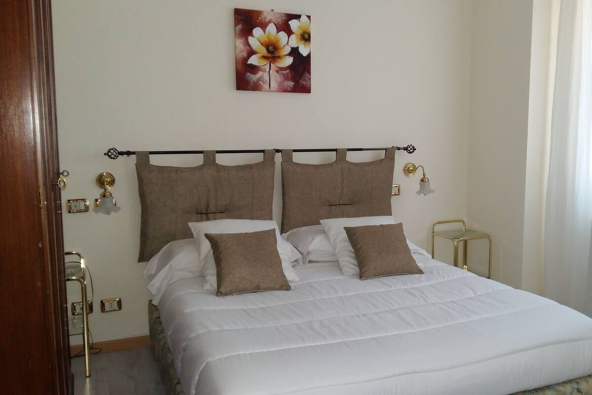 Una cama bien hecha con ropa blanca y dos almohadas marrones, un cabecero de madera, una mesita de noche con una lámpara, y una obra de arte colgada en la pared con una flor.