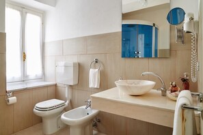 Une salle de bain moderne comprenant des toilettes, un bidet, un lavabo avec un receveur en marbre, un porte-serviettes, un miroir et une fenêtre avec des rideaux.