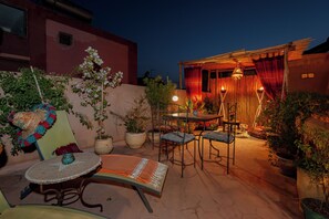 Un patio al aire libre con una mesa redonda, dos sillas y una pequeña mesa con un adorno. Hay plantas en macetas y un edificio al fondo.