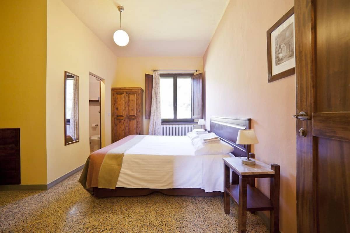 A hotel room with a large bed, a bedside table with a lamp, a window with curtains, and a door with a framed picture.