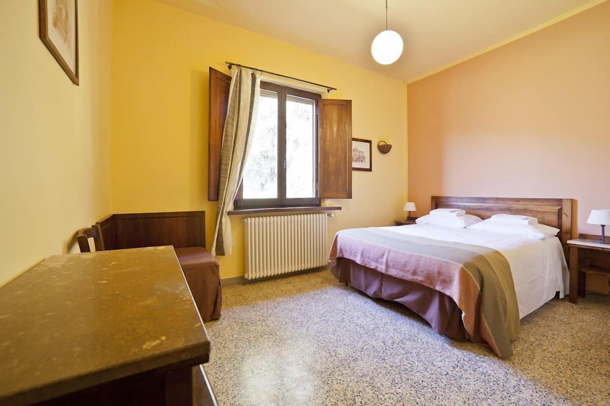 A bedroom with a bed, a table, a chair, a window with curtains, and a radiator.