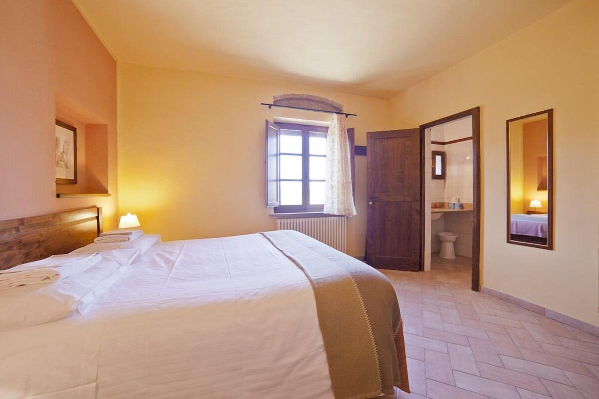 A hotel room with a bed, a window with curtains, a wooden door, a bathroom, and a mirror.