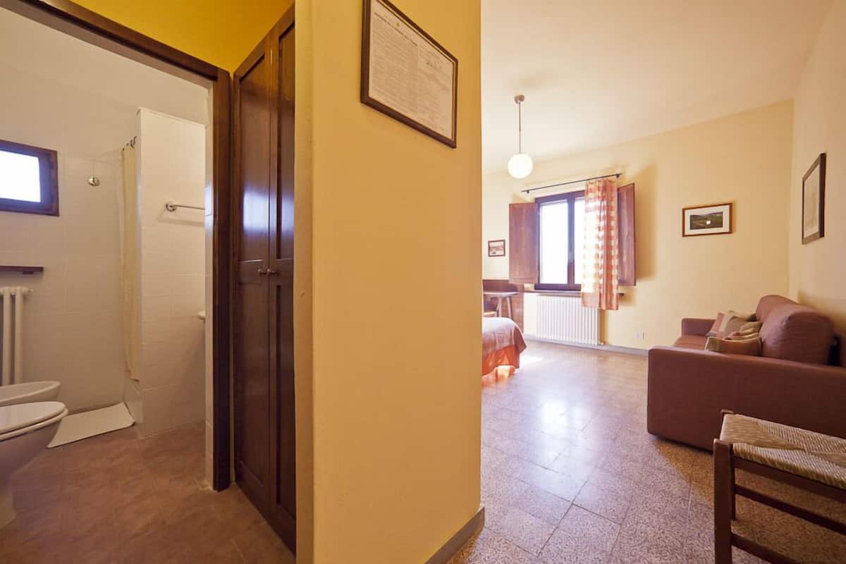 A hallway leading to a bathroom with a toilet and shower, a living area with a sofa and a chair, and a dining area with a table and chairs.