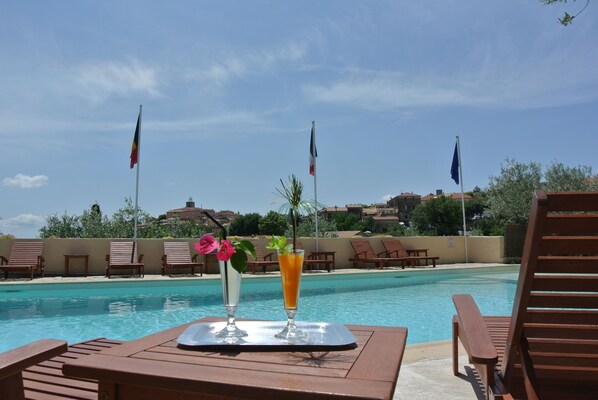 Zona de piscina con sillones de descanso y una mesa con bebidas. Se ven banderas al fondo.