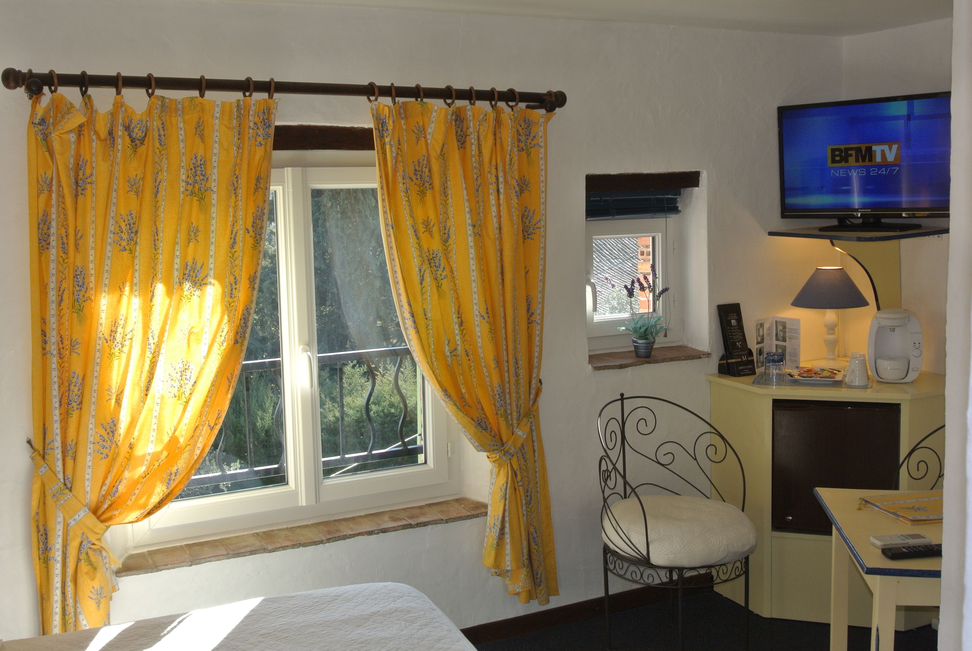 Habitación de hotel con cama, escritorio con lámpara, televisor mostrando 'BFMTV NEWS 24/7' y cortinas de estampado floral.